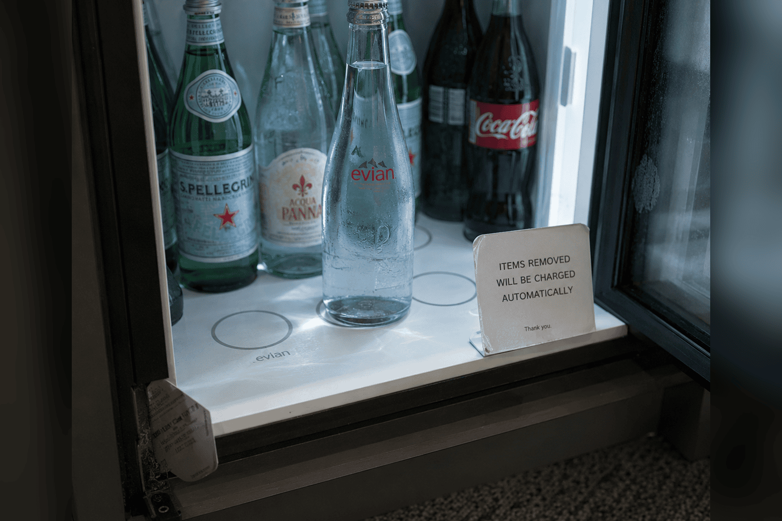 A minibar shelf where one water bottle stands off its sensor ring, beside a sign: removed items are charged automatically.