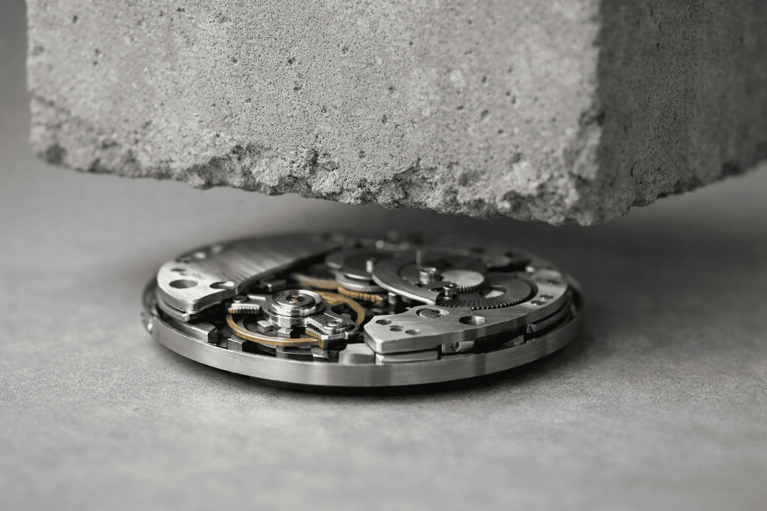 A heavy concrete block hovers just above an exposed mechanical watch movement on a grey stone surface.
